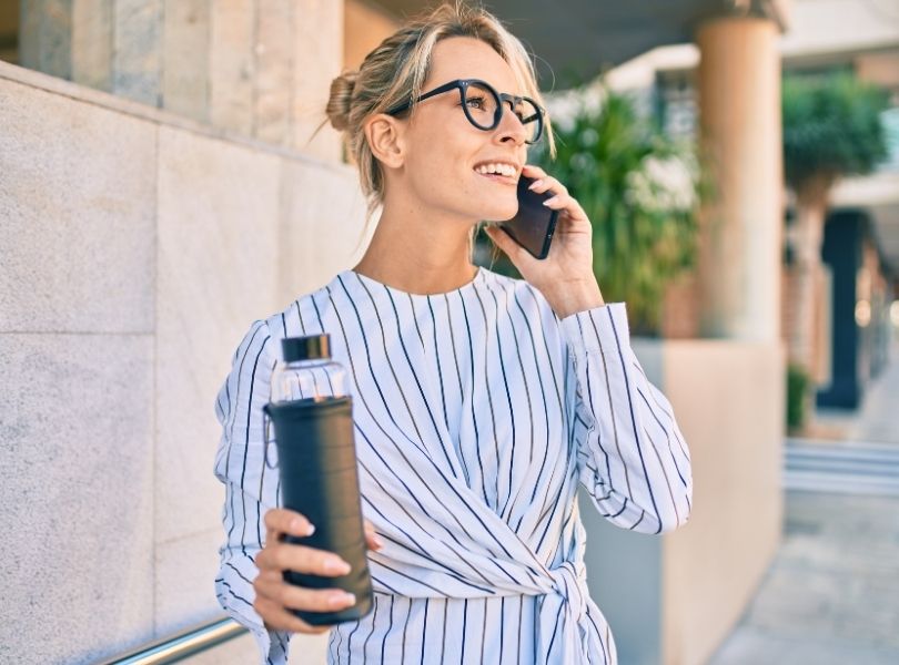 A woman walking on a sidewalk with coffee and speaking on a telephone.