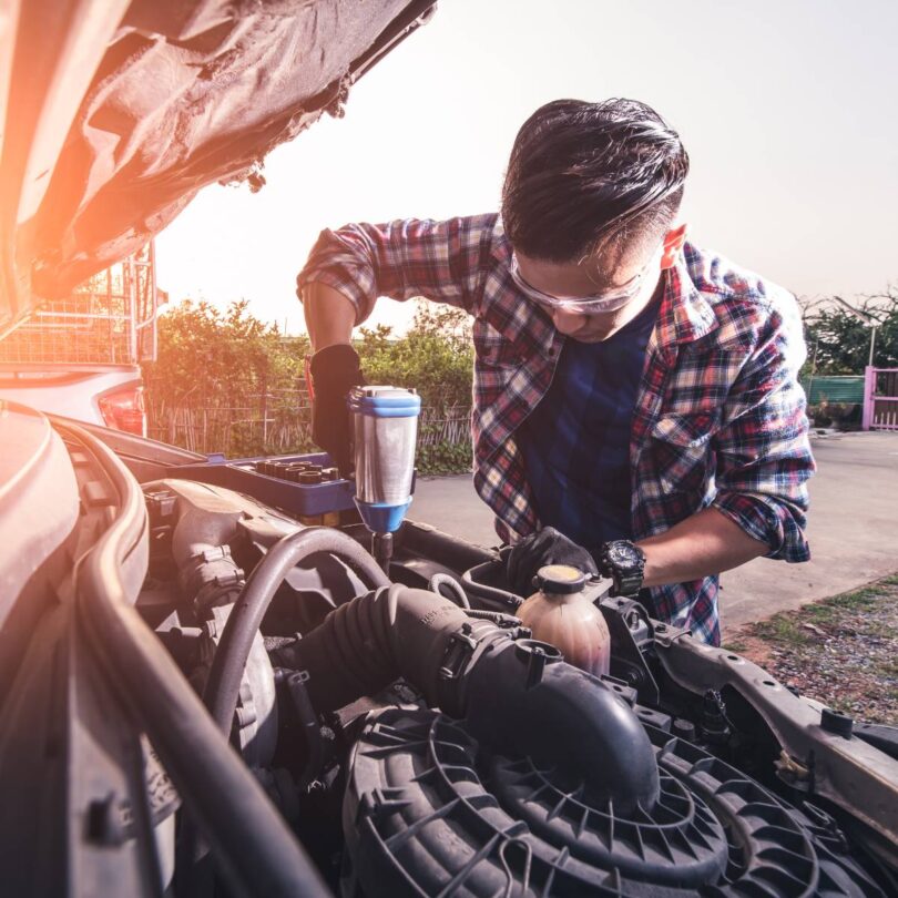 A guy wearing protective goggles and a flannel shirt is modifying or repairing a vehicle's engine in a car junkyard.