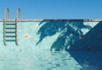An empty swimming pool with turquoise tiles, a small step ladder, and the shadow of a palm tree on a beautiful, clear day.