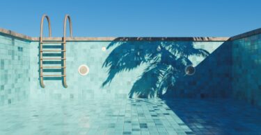An empty swimming pool with turquoise tiles, a small step ladder, and the shadow of a palm tree on a beautiful, clear day.
