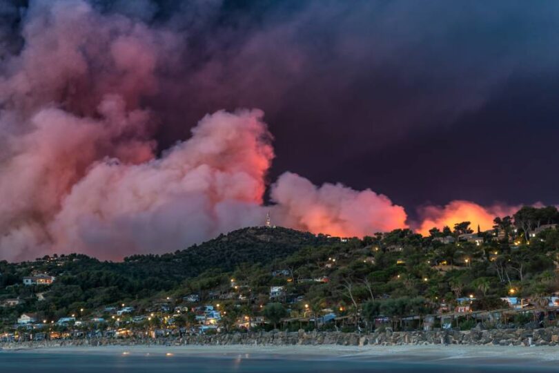 A wildfire engulfing a beach city and filling the air with smoke, smog, ash, and flame, destroying homes.
