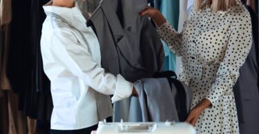Two women, one in a polka dot dress and the other in a white top, are smiling and putting together an outfit in a store.