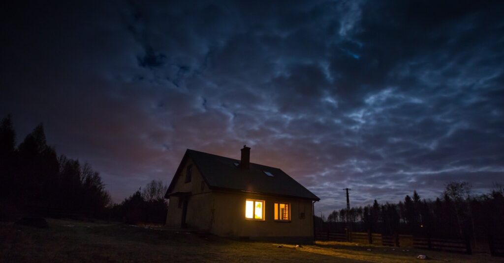 A home lit up only by a light in a window sits alone in the dark with trees in the background and dark clouds in the sky.
