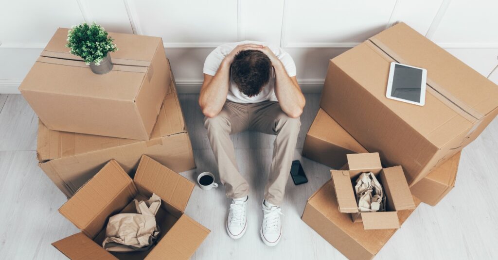 A bird's-eye view of a man sitting with his head in his knees. Several half-packed boxes are surrounding him.