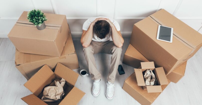 A bird's-eye view of a man sitting with his head in his knees. Several half-packed boxes are surrounding him.
