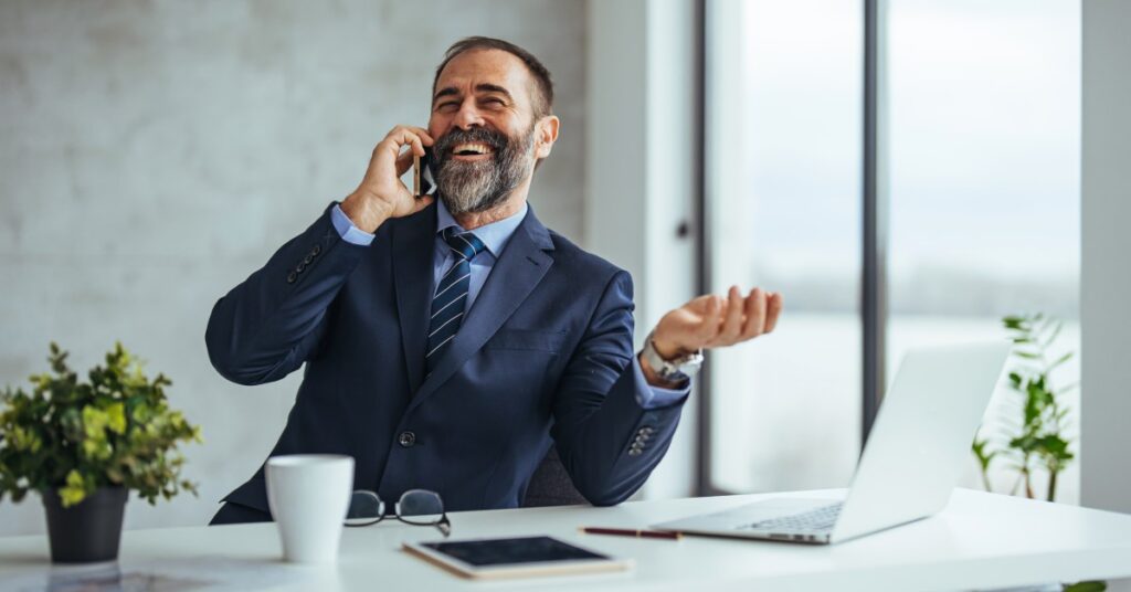 A businessman wearing a blue suit smiles down at his phone. He's sitting at a desk with a laptop computer, tablet, and notes.