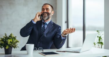 A businessman wearing a blue suit smiles down at his phone. He's sitting at a desk with a laptop computer, tablet, and notes.