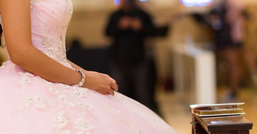 A young woman wearing a pale pink Quinceañera dress that’s embellished with rhinestones on the bodice and skirt.