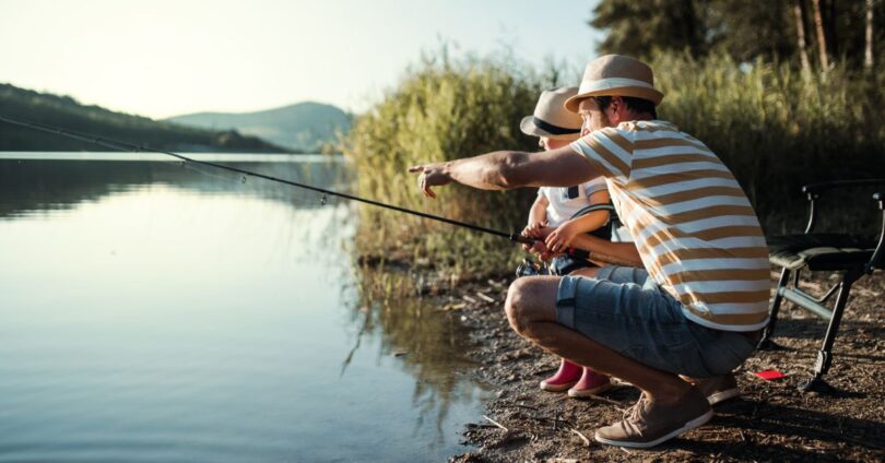 A father and young son, both wearing fedoras, crouching by the bank of a lake with a fishing rod, trying to catch some fish.