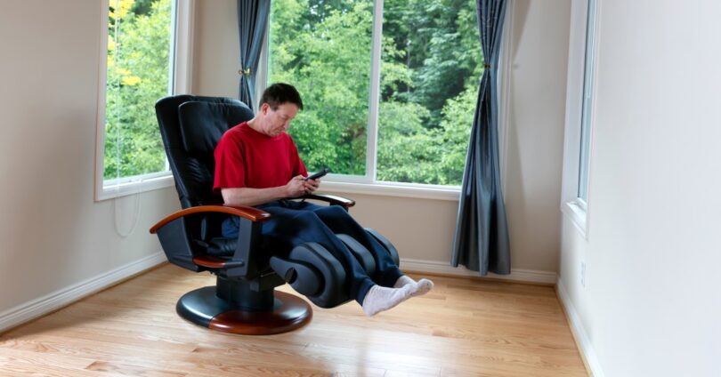 A homeowner using his massage chair at home. He's wearing comfortable clothing and using the remote to start his session.