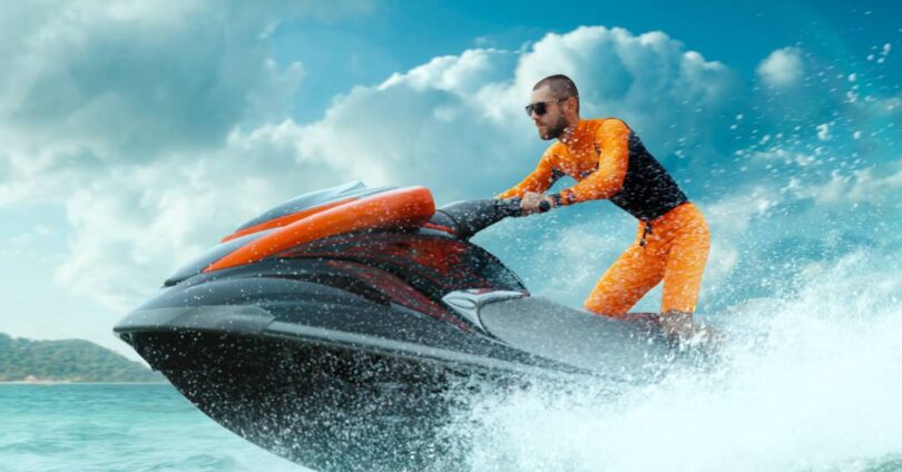 A person wearing a jet ski suit rides on a sleek red and black jet ski on the water, leaving big waves in its wake.