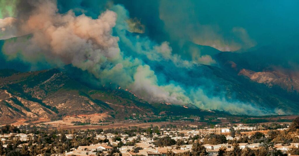 A wildfire blazes in a forest on a mountain as the smoke billows towards the sky. A town appears at the bottom of the mountain.