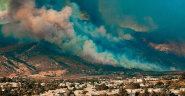 A wildfire blazes in a forest on a mountain as the smoke billows towards the sky. A town appears at the bottom of the mountain.