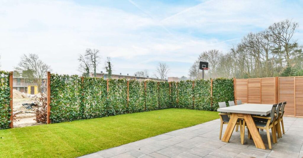 A backyard terrace has a table with some chairs on some concrete, next to a grassy area framed by decorative wall plants.