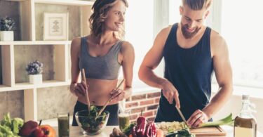 Woman and man wearing workout clothing inside a kitchen chopping vegetables and mixing greens while smiling.