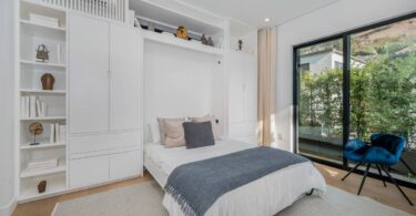A bedroom with a Murphy bed. The bed has a white duvet with gray and tan pillows. A gray throw blanket is on the bed, too.
