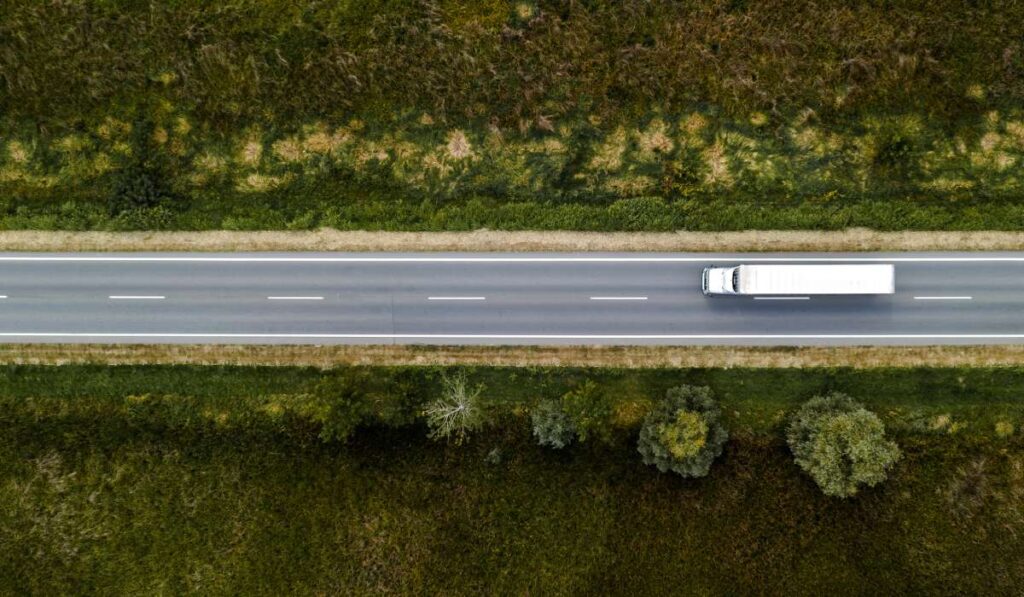 An aerial view of a large white semi-truck driving down a highway with no other cars on the road. Trees and grass line the road.
