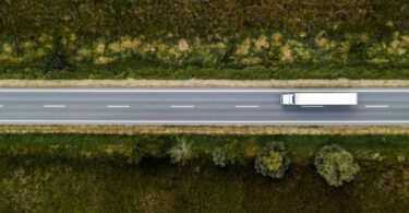 An aerial view of a large white semi-truck driving down a highway with no other cars on the road. Trees and grass line the road.