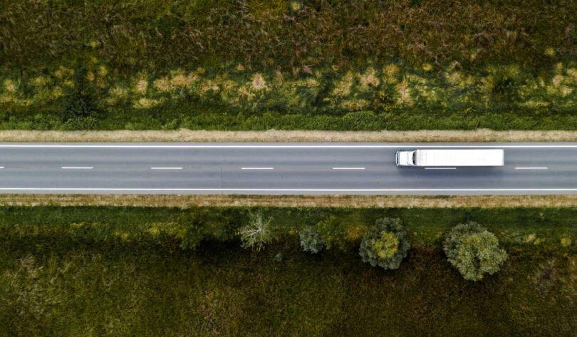 An aerial view of a large white semi-truck driving down a highway with no other cars on the road. Trees and grass line the road.