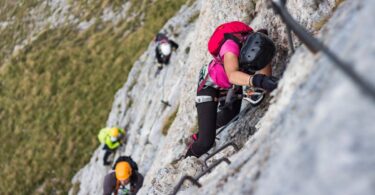 A group of climbers ascend a cliff face using ropes and mounted holds. The sloping face of a mountain is seen below.