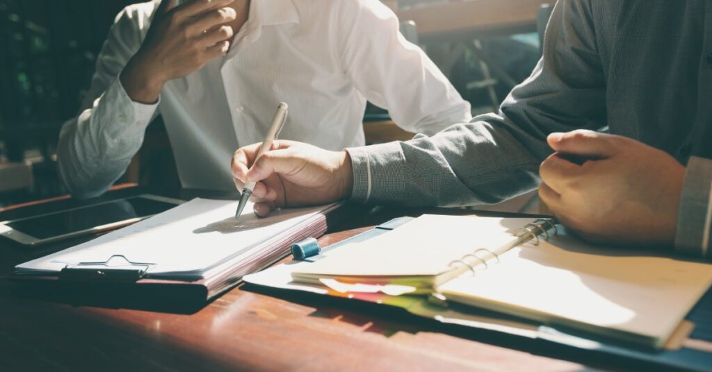 Two men sitting at a desk. One is writing on a form attached to a clipboard with a pen while an open binder sits in front of him.