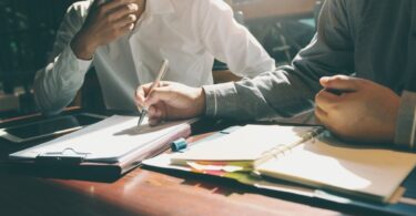 Two men sitting at a desk. One is writing on a form attached to a clipboard with a pen while an open binder sits in front of him.