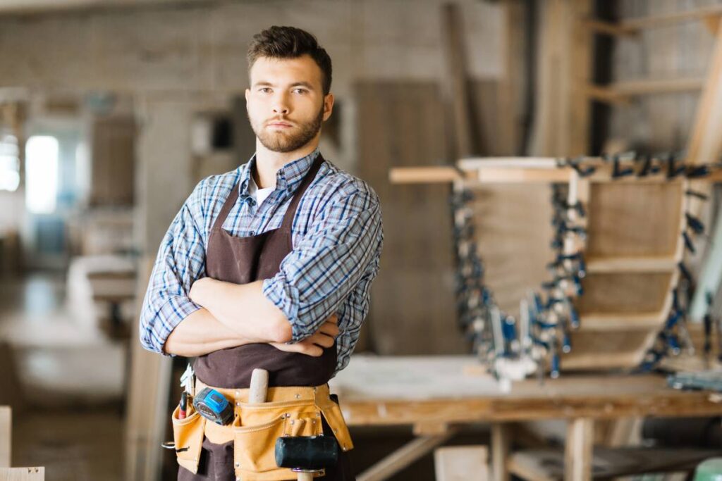 A woodworker stands in his shop, wearing a leather toolbelt and a brown apron. He has a serious look on his face.