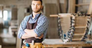 A woodworker stands in his shop, wearing a leather toolbelt and a brown apron. He has a serious look on his face.