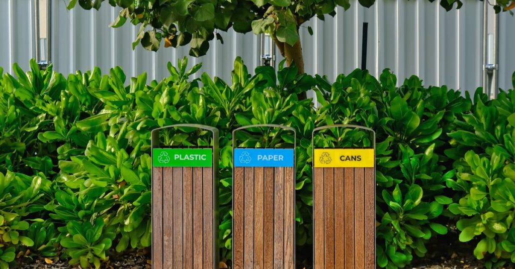 A set of plastic, paper, and cans waste baskets. These bins are sitting in front of a vibrant tree and bushes.