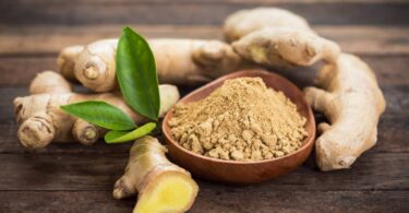 Multiple ginger roots surround a small wooden bowl filled with ground ginger. The items lie on a wooden table.