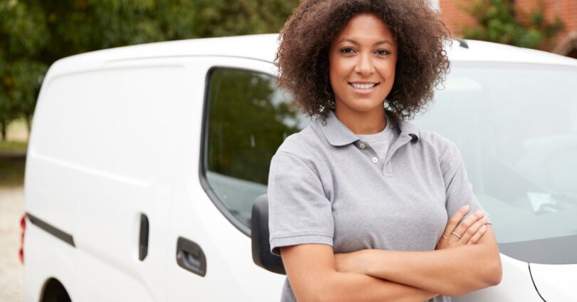 A woman wearing a grey polo with her arms crossed stands in front of a parked white company vehicle.