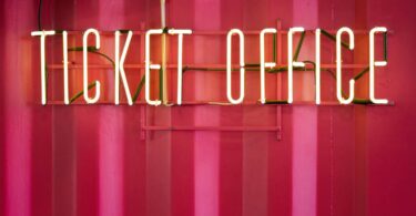A bright neon sign reading "Ticket Office" is hanging on the side of a bright red shipping container.