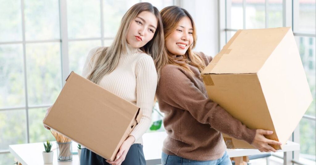 Two young Asian women smiling and posing with two large moving cardboard boxes in their new apartment.