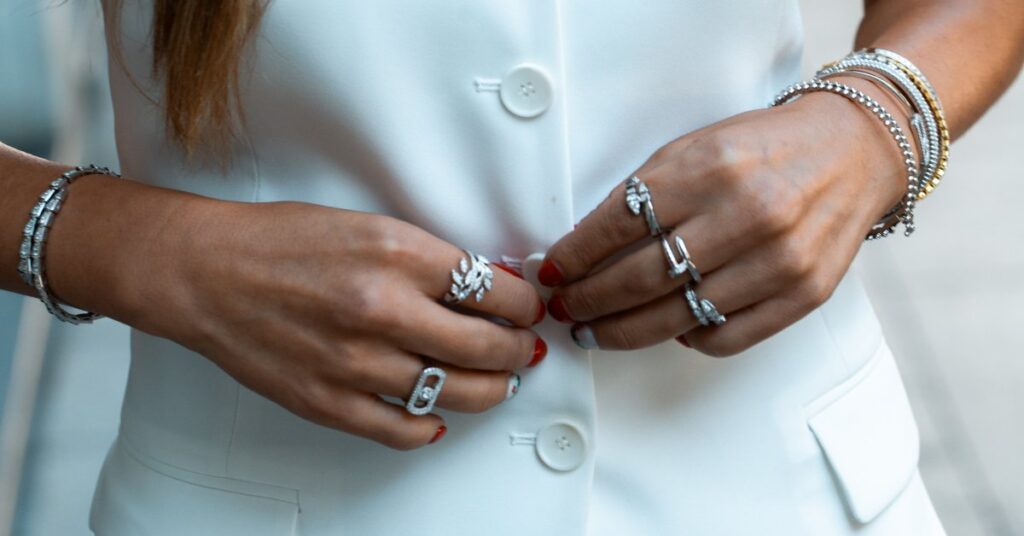A close-up of a young woman in a white vest wearing various mixed-metal bracelets, rings, and necklaces that she layered.