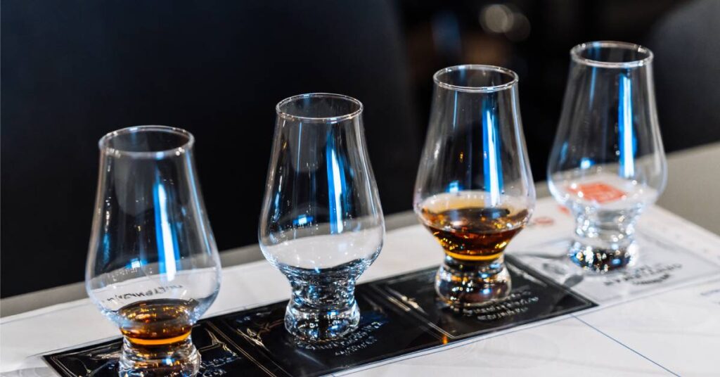 Four clear glasses lined up in a row on a bar countertop. Two out of the four glasses have a light brown liquid in them.