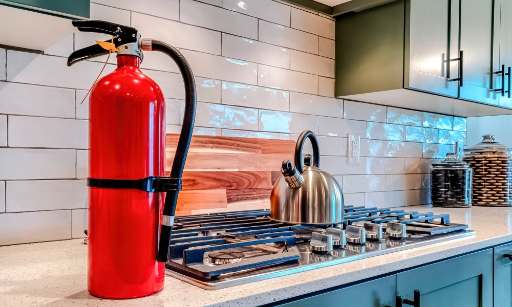 A red fire extinguisher is sitting on a kitchen counter next to a cooktop with a stainless steel kettle on the burner.