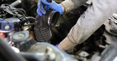 A person wearing blue gloves holds a car's air intake hose near an engine bay, surrounded by various engine components.