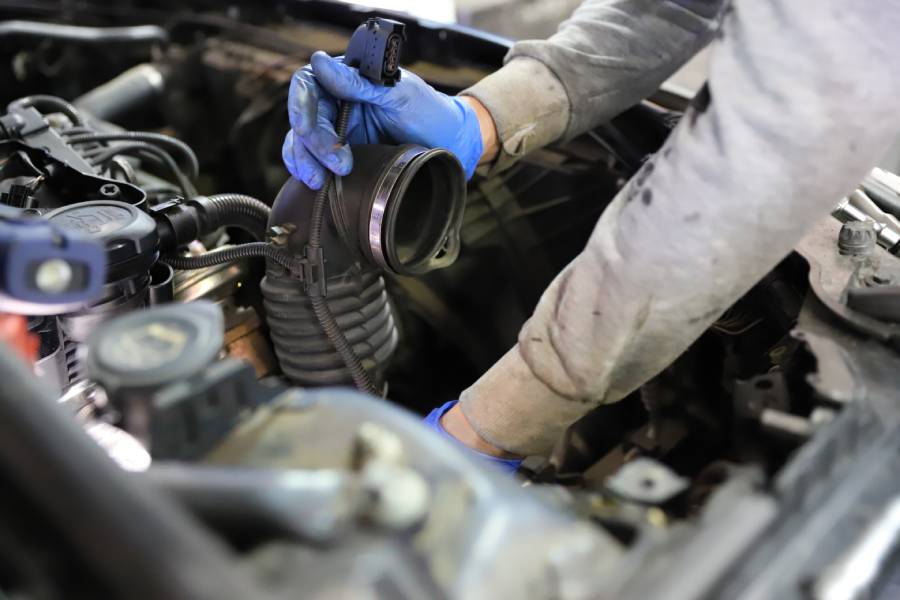 A person wearing blue gloves holds a car's air intake hose near an engine bay, surrounded by various engine components.