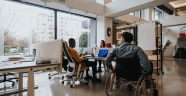 A modern-looking office with various workstations. Two people sit in office chairs while a man uses a wheelchair.