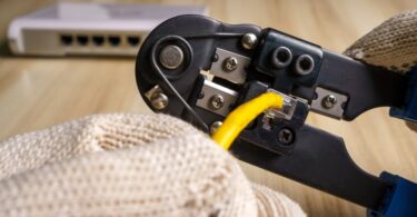 A close-up view shows a hand wearing a tan glove using a black crimper to crimp yellow wires together.