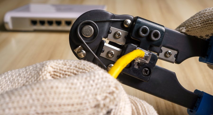 A close-up view shows a hand wearing a tan glove using a black crimper to crimp yellow wires together.