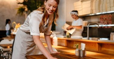 A young woman wearing a gray apron uses an orange cloth to wipe down a wooden table in a restaurant.