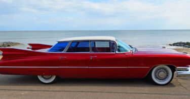 A classic red and white vintage 1950s 4-door Cadillac with wire wheels that is parked on a road near an ocean.