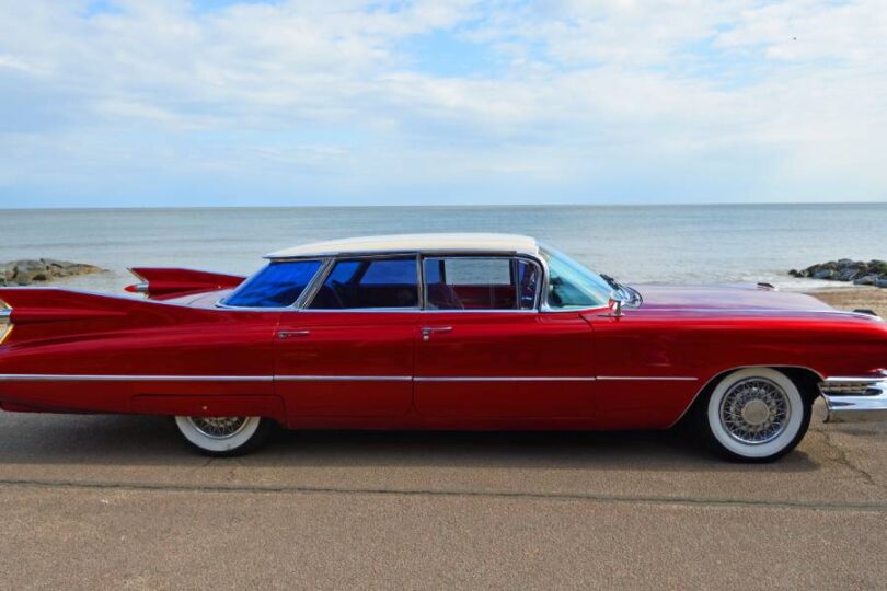 A classic red and white vintage 1950s 4-door Cadillac with wire wheels that is parked on a road near an ocean.