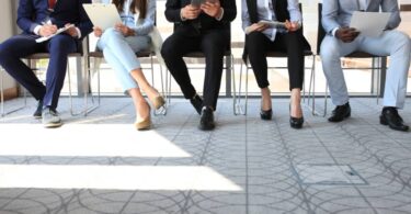 A row of five people wearing business casual attire sit in a row of chairs while holding clipboards and papers.