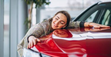 A woman happily hugs the hood of a red car in a car dealership. A potted tree stands behind her in the background.