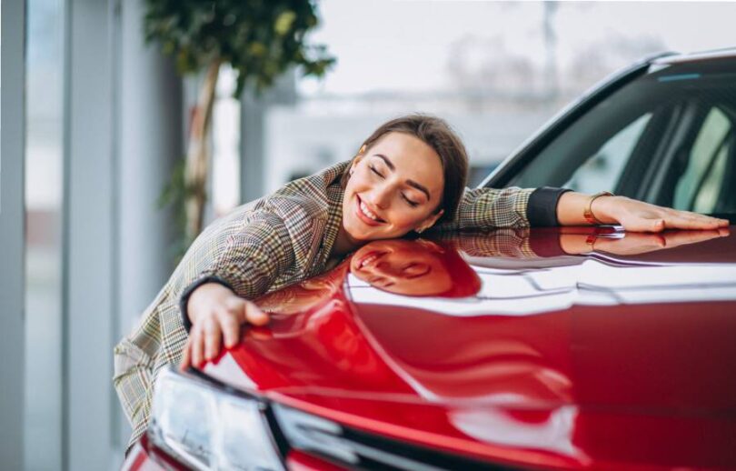 A woman happily hugs the hood of a red car in a car dealership. A potted tree stands behind her in the background.