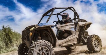 A person wearing protective gear rides an off-road vehicle on a dirt road with dust flying around the tires.