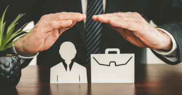 A businessperson in a black blazer and tie with their hands over a person and briefcase wooden figurine.