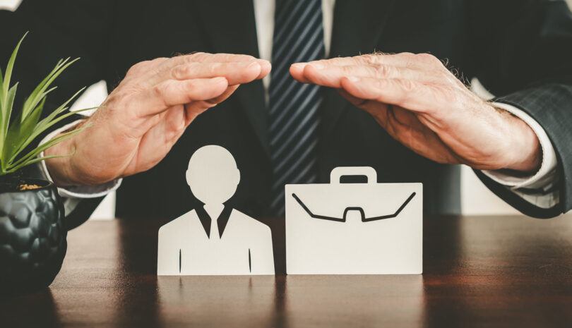 A businessperson in a black blazer and tie with their hands over a person and briefcase wooden figurine.